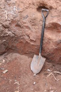 Image of a shovel in dirt beside a rock reddish clay covered ground with black and silver shovel